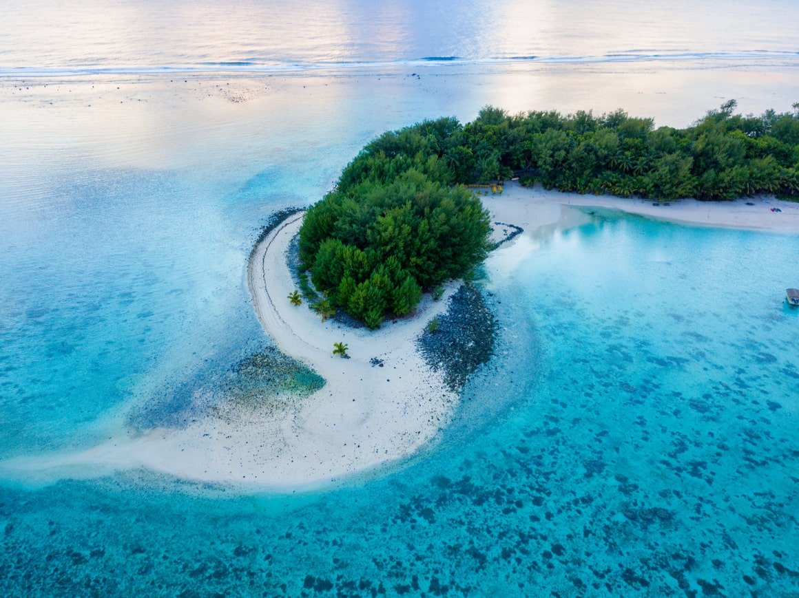 Very clear, blue water is broken up by a peninsula with greenery on it in the Cook Islands. 