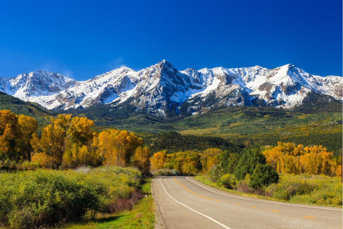 Snow capped mountains and autumn trees are in the distance. A straight road with no cars on it is in the foreground. 