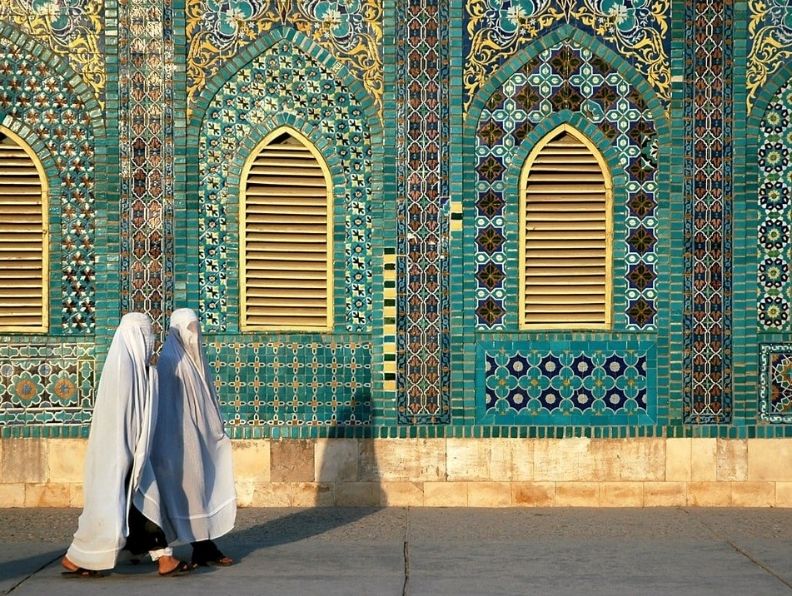 two women in white burqas walking past a turquoise colored shrine in afghanistan