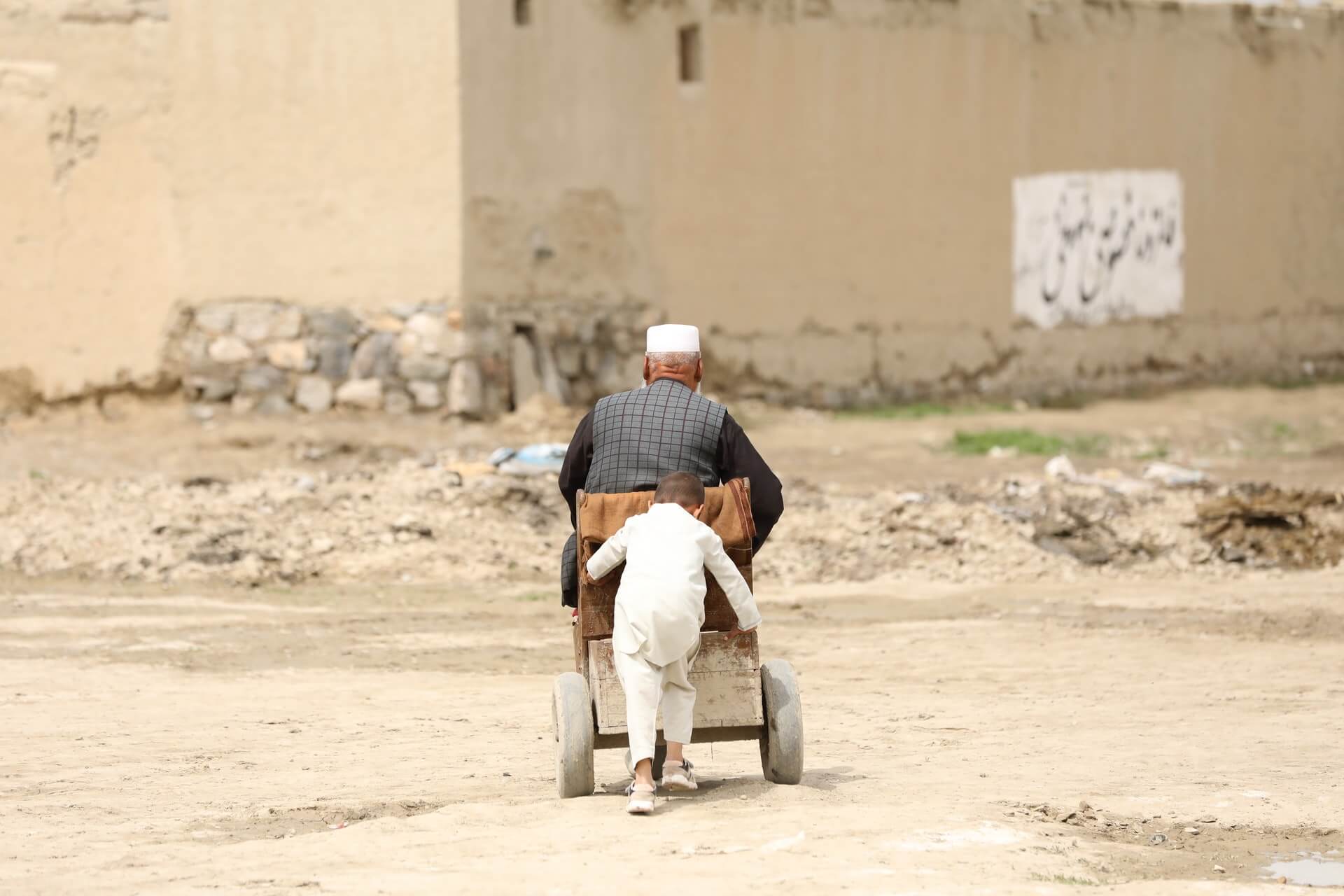 A small Afghan boy pushes his grandfather down a dusty road in a cart.