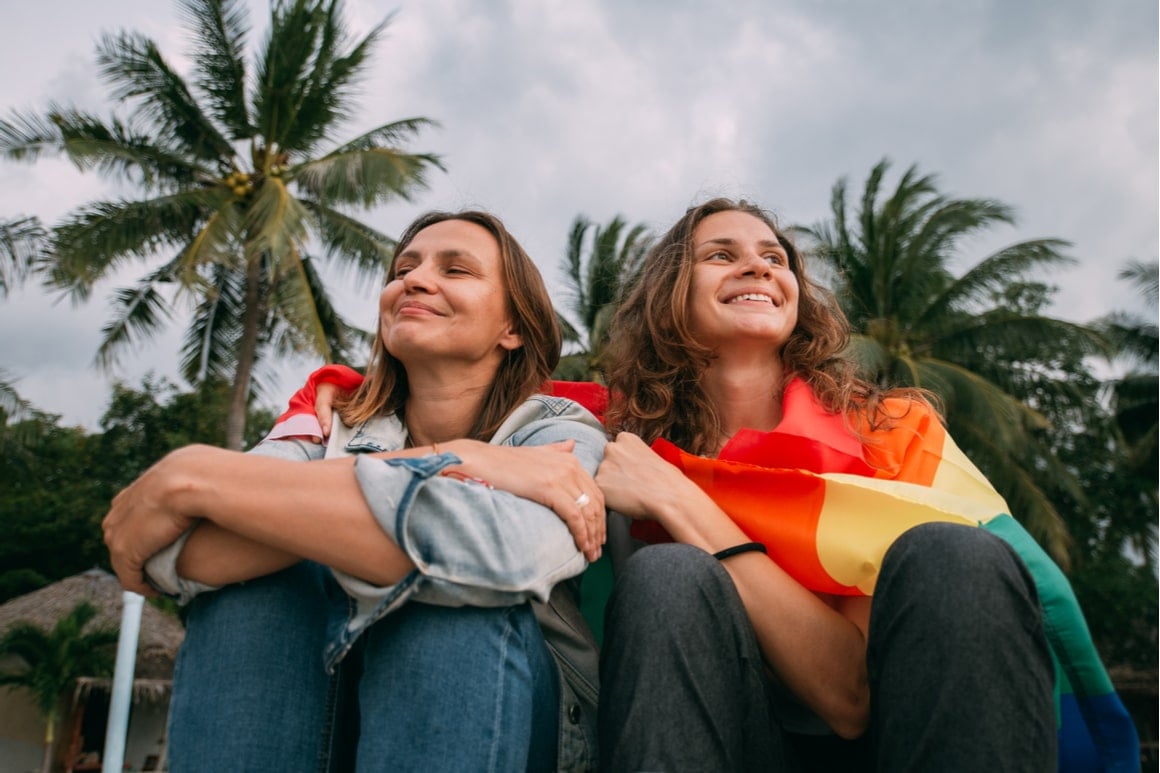 Two girls sit next to each other with the LGBTQ pride flag wrapped around them. 