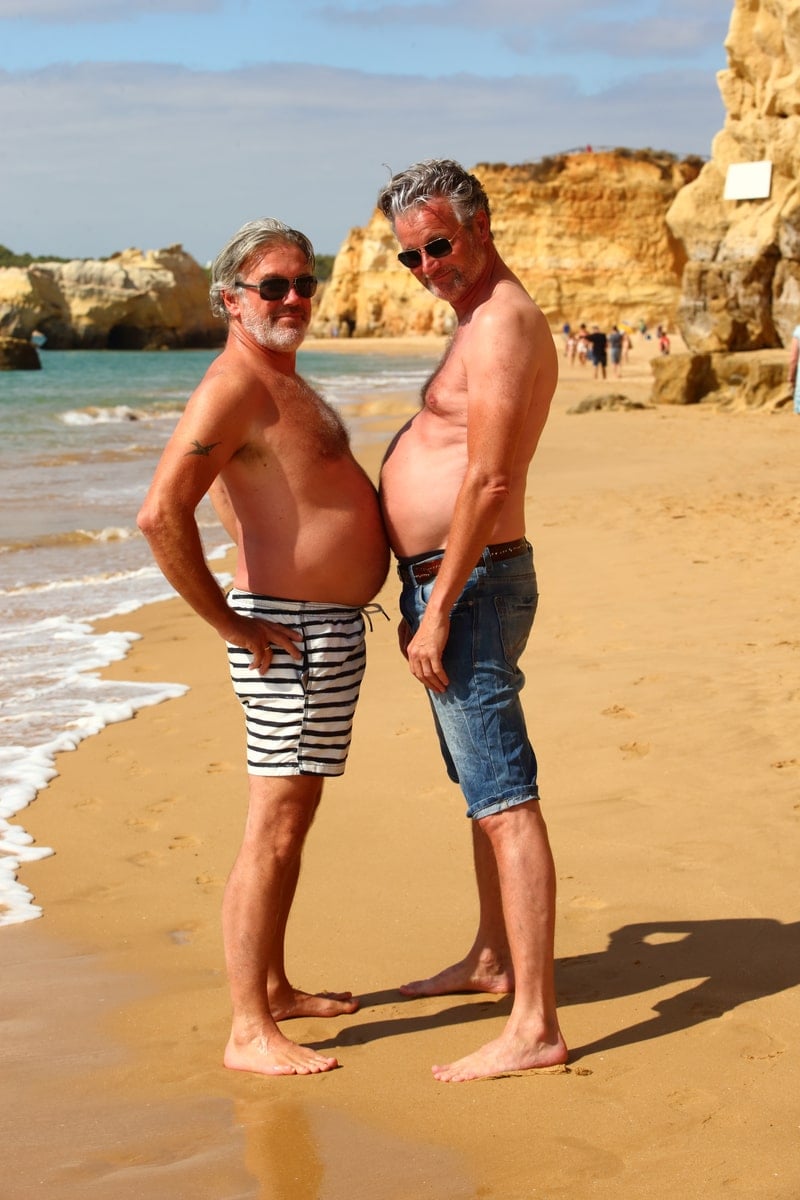Two men show off their beer bellies while travelling gay together.