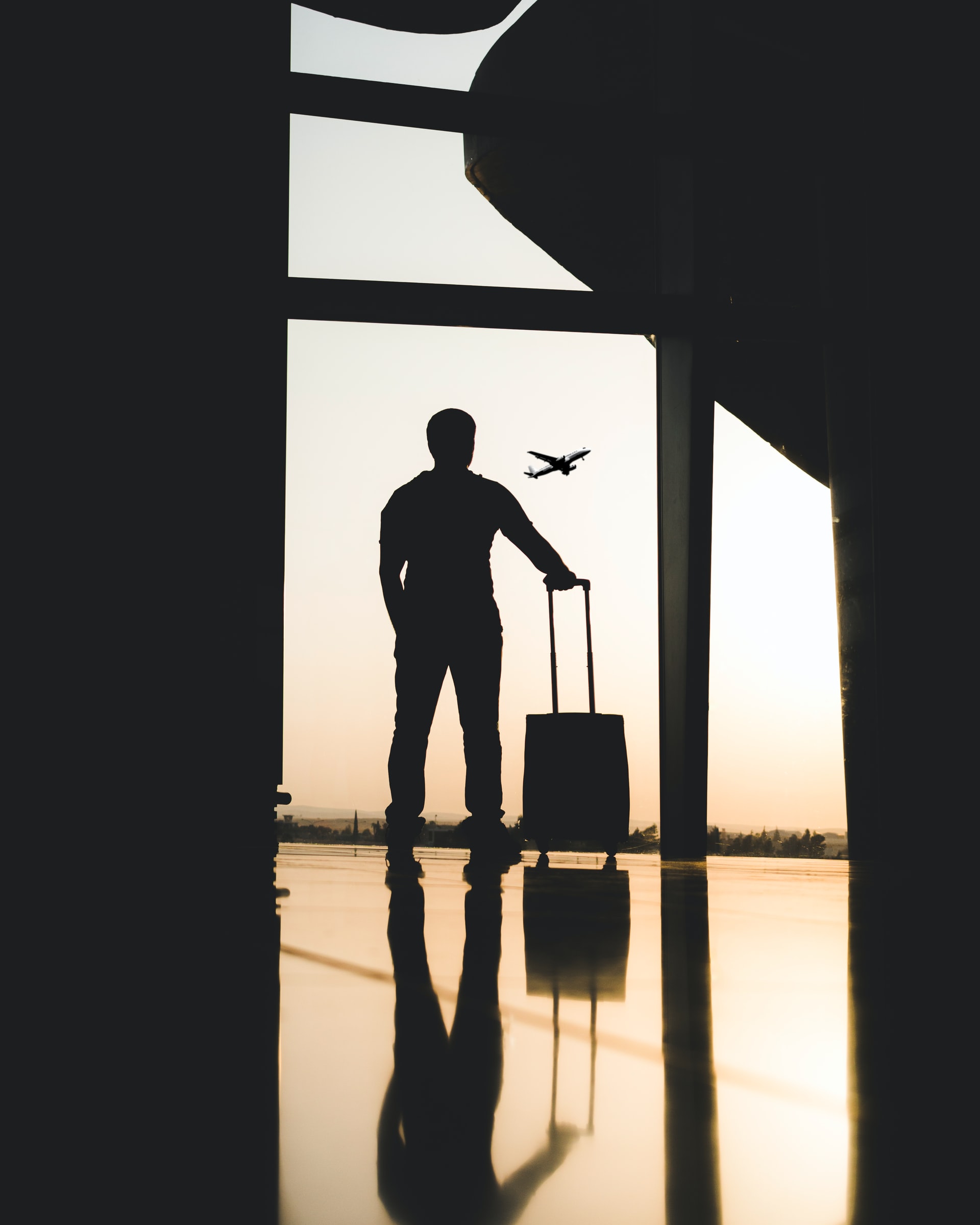 man with a suitcase silhouetted against an airport window watching a plane