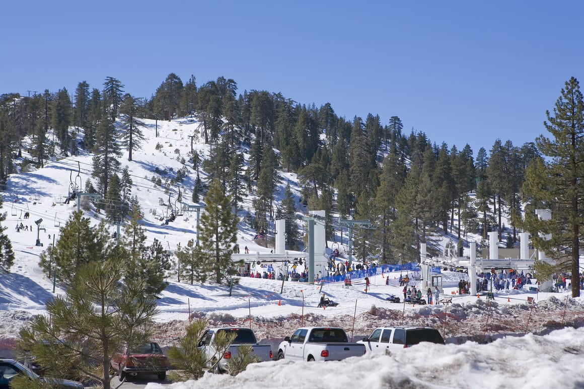 A picture from the parking lot at Big Bear Mountain Ski Resort showing a chairlift  on a background of snowy pine covered mountains and people entering the resort