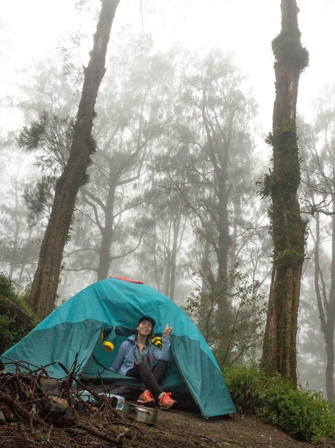 Hiker sitting in front of a blue tent in a misty forest on a mountain in Bali.