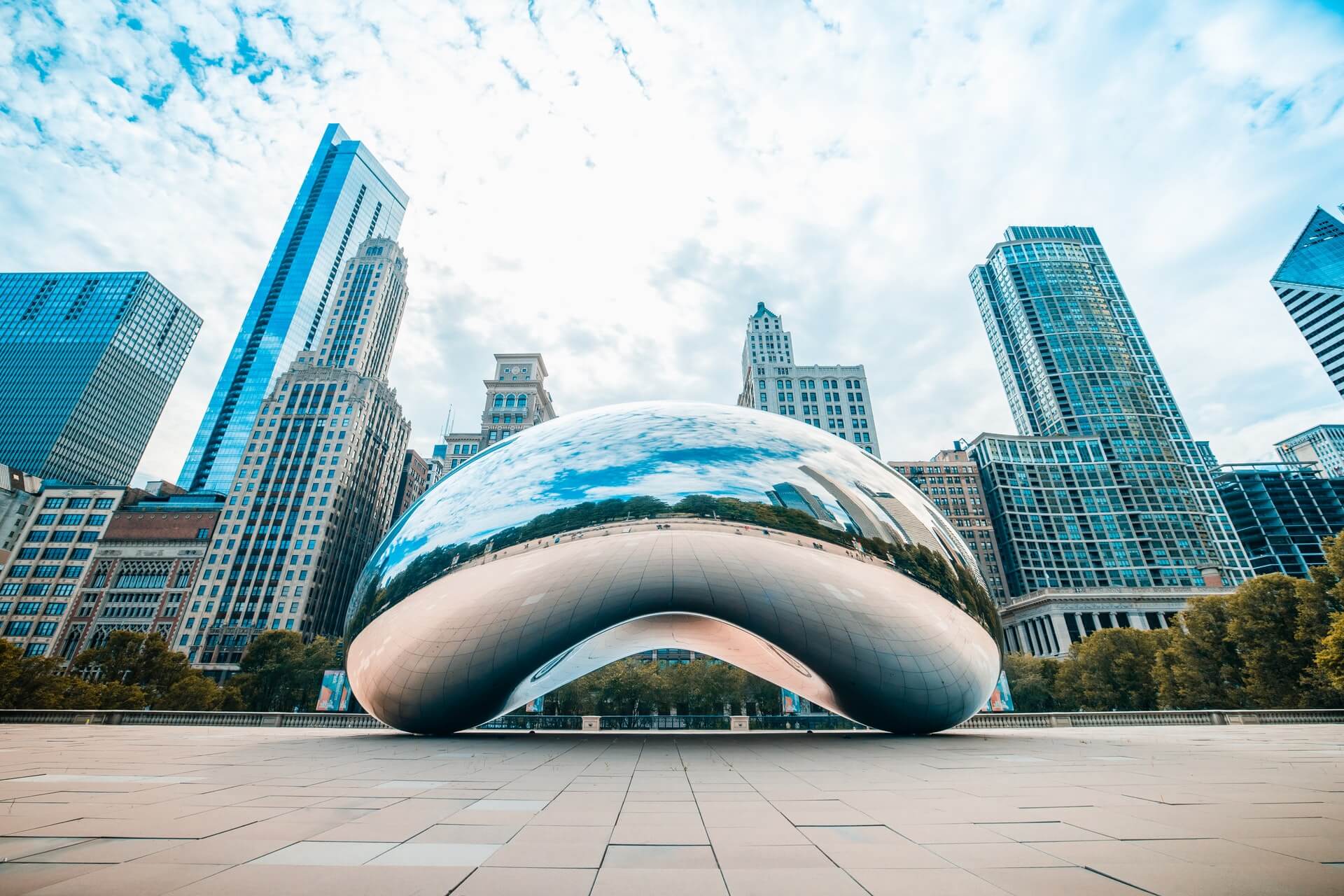silver massive reflective bean in front of high rise buildings with windows traveling in chicago