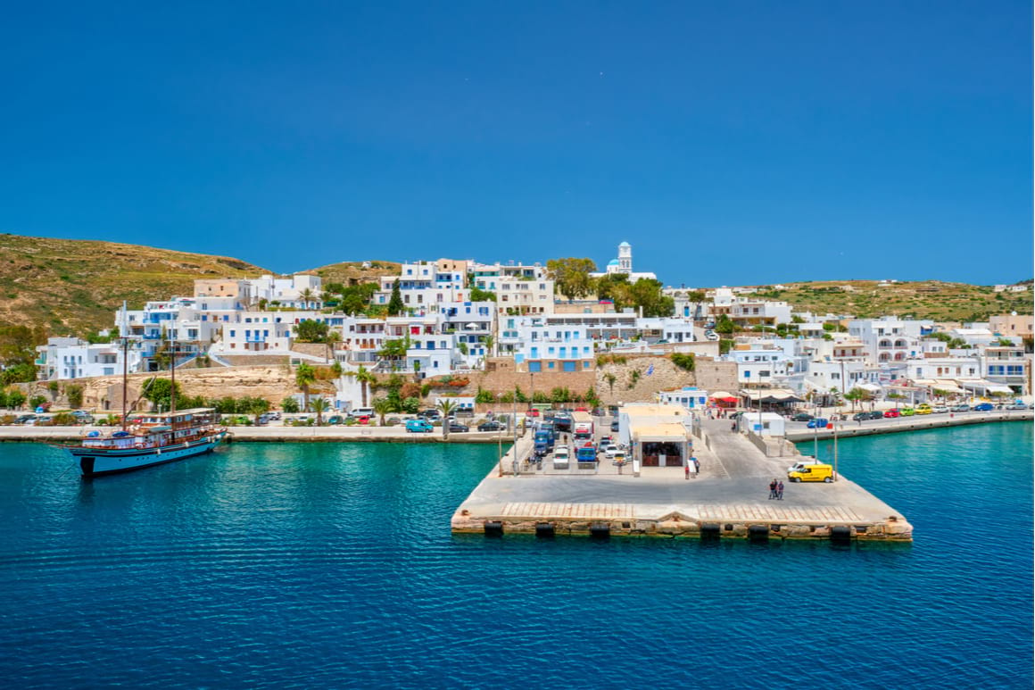 View of Adamas from the water. The sky and sea are super blue. Photo shows the port area and the white and coloured houses on the hill behind.