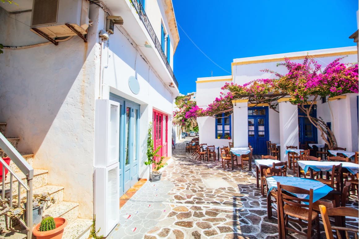 In Plaka town. Cobbled street, tables outside and big flowers on the white buildings.