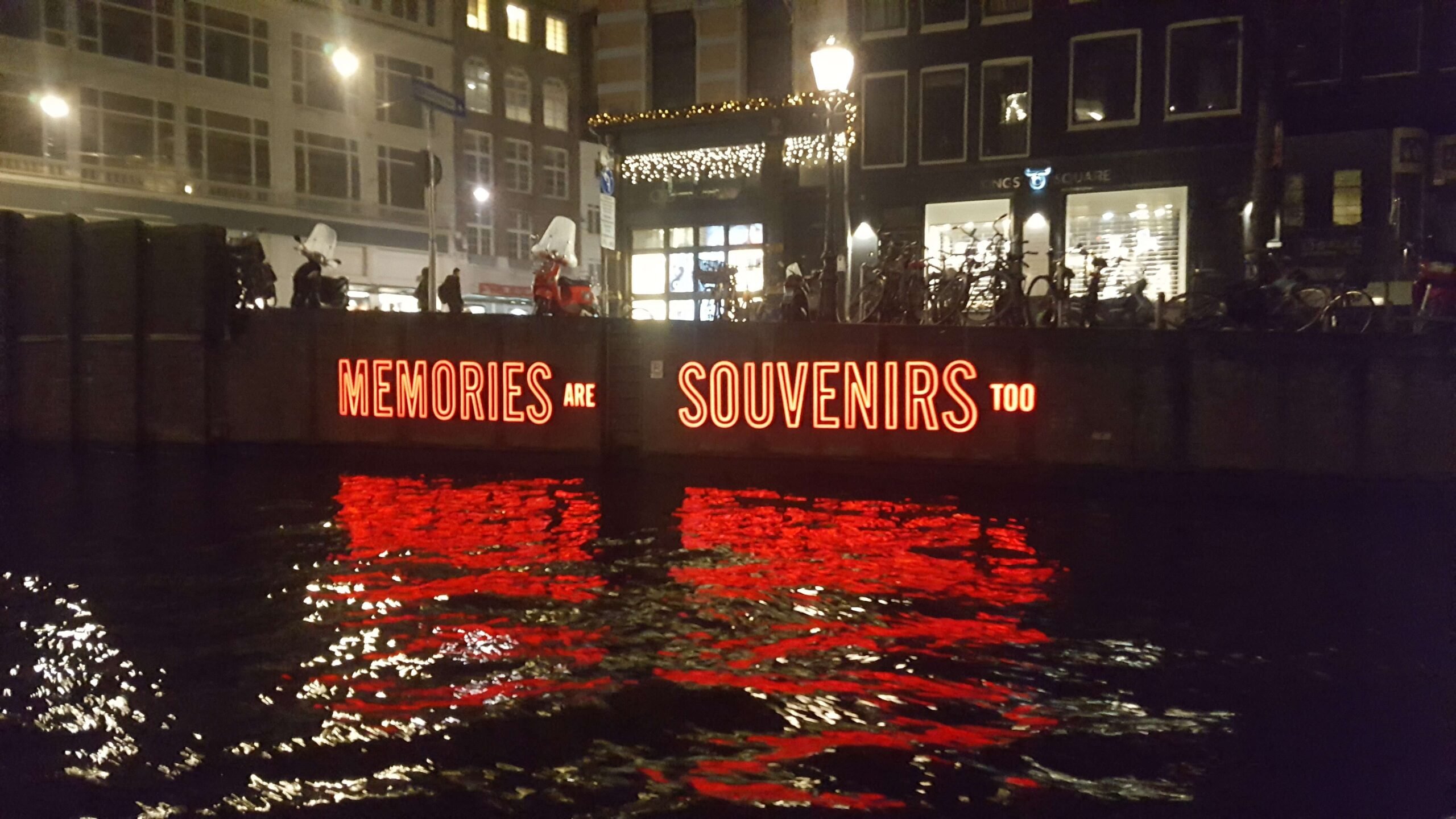 Red neon light art reading "memories are souvenirs too" on the side of the canal at night