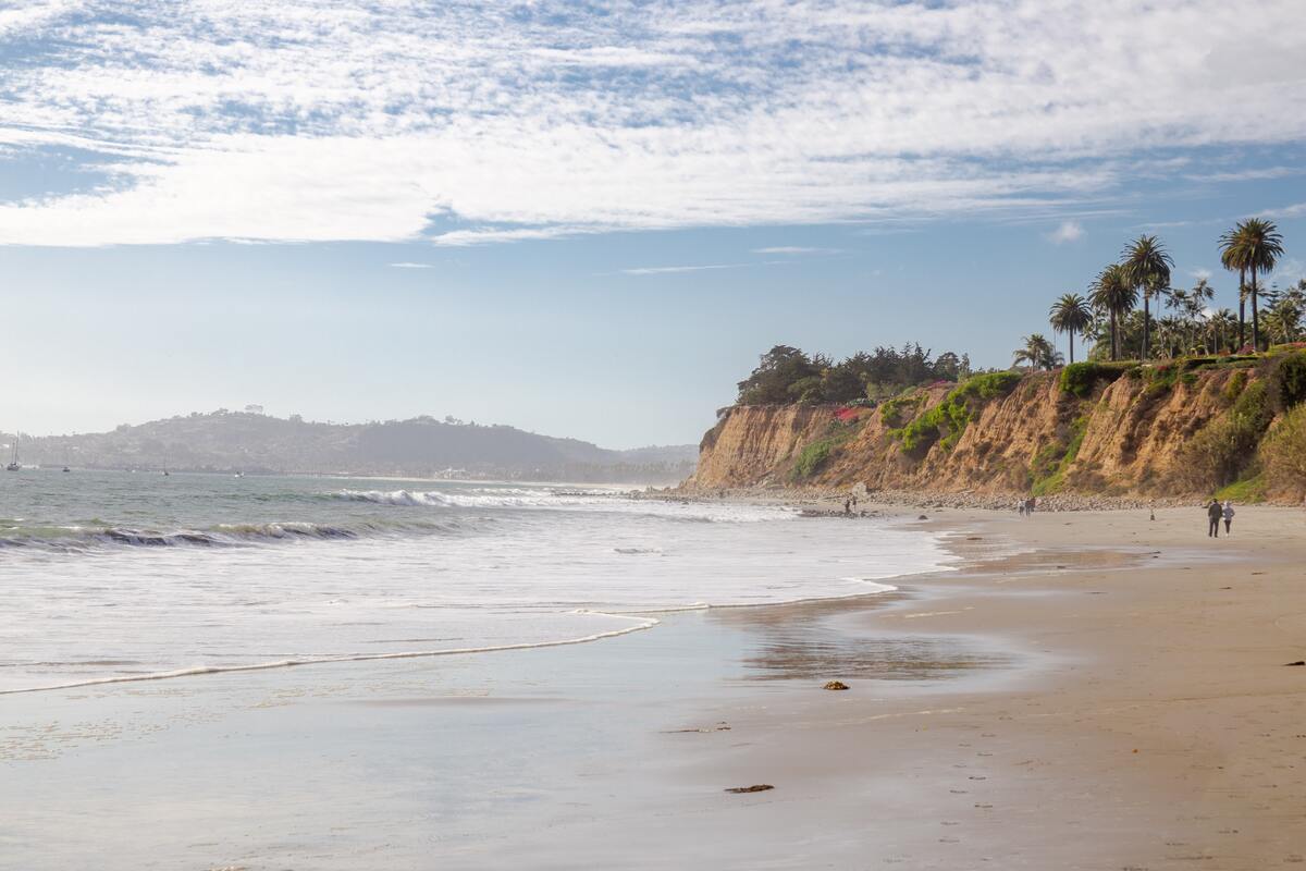 people walking on a beach with small tree lined mountain cliffs rising up behind and in front of them in santa barbara