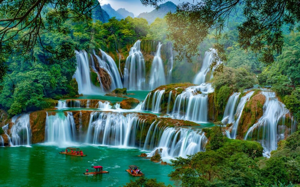 tourists in orange boats on the pool of a wide emerald waterfall that's shared between vietnam and china