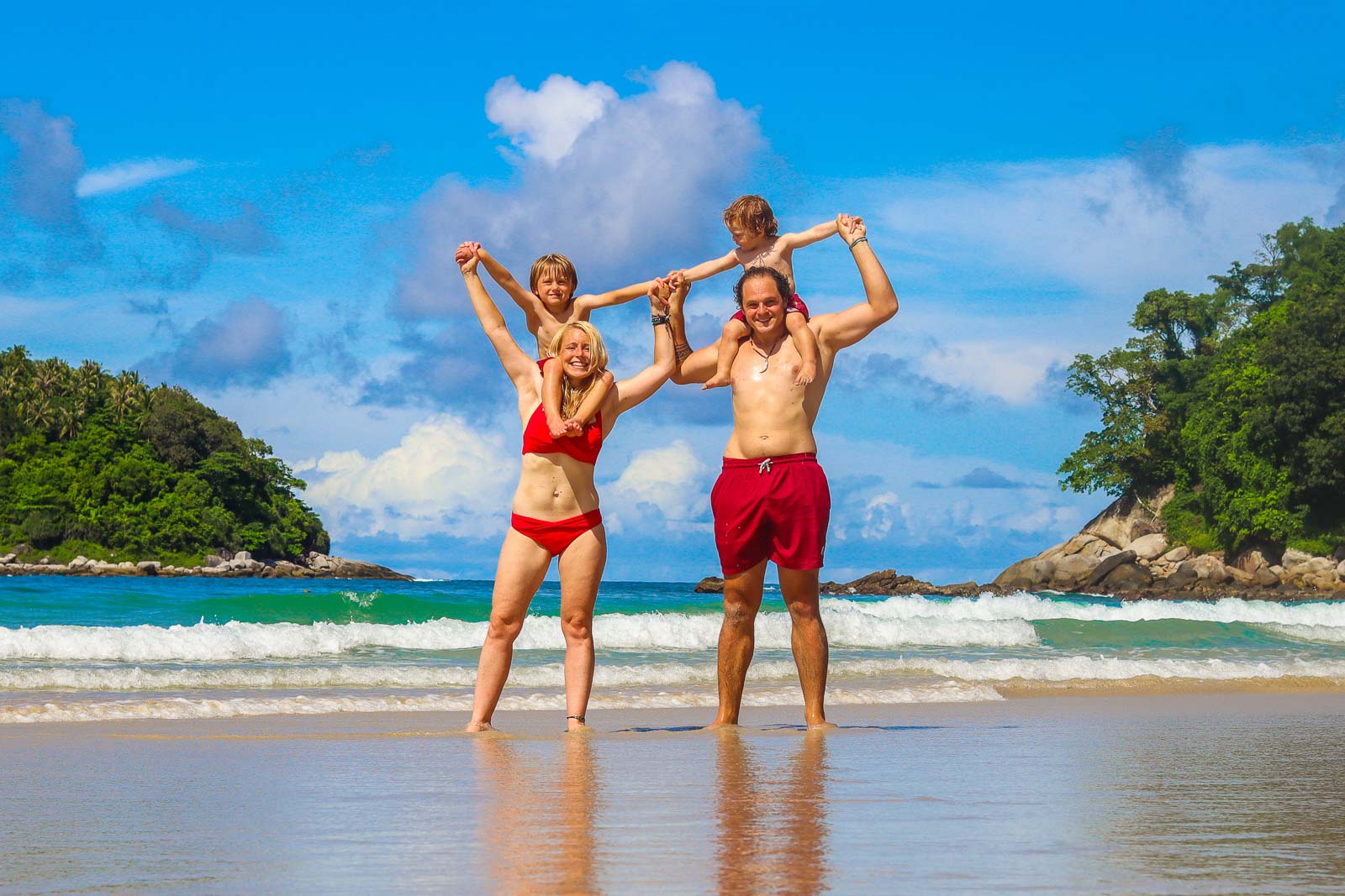 couple standing on a stunning tropical beach with one kid on each of their shoulders