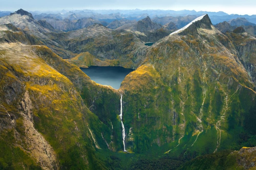 a white fall tumbling down a green colored mountain in new zeland beneath a circular blue lake