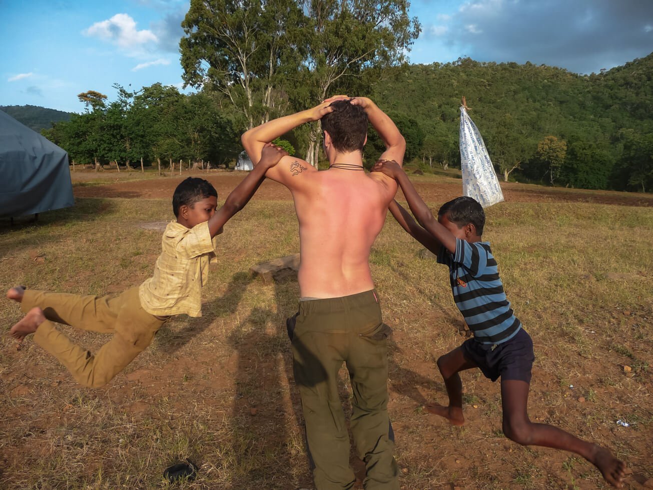 shirtless man volunteering in rural india with two kids swinging on his arms