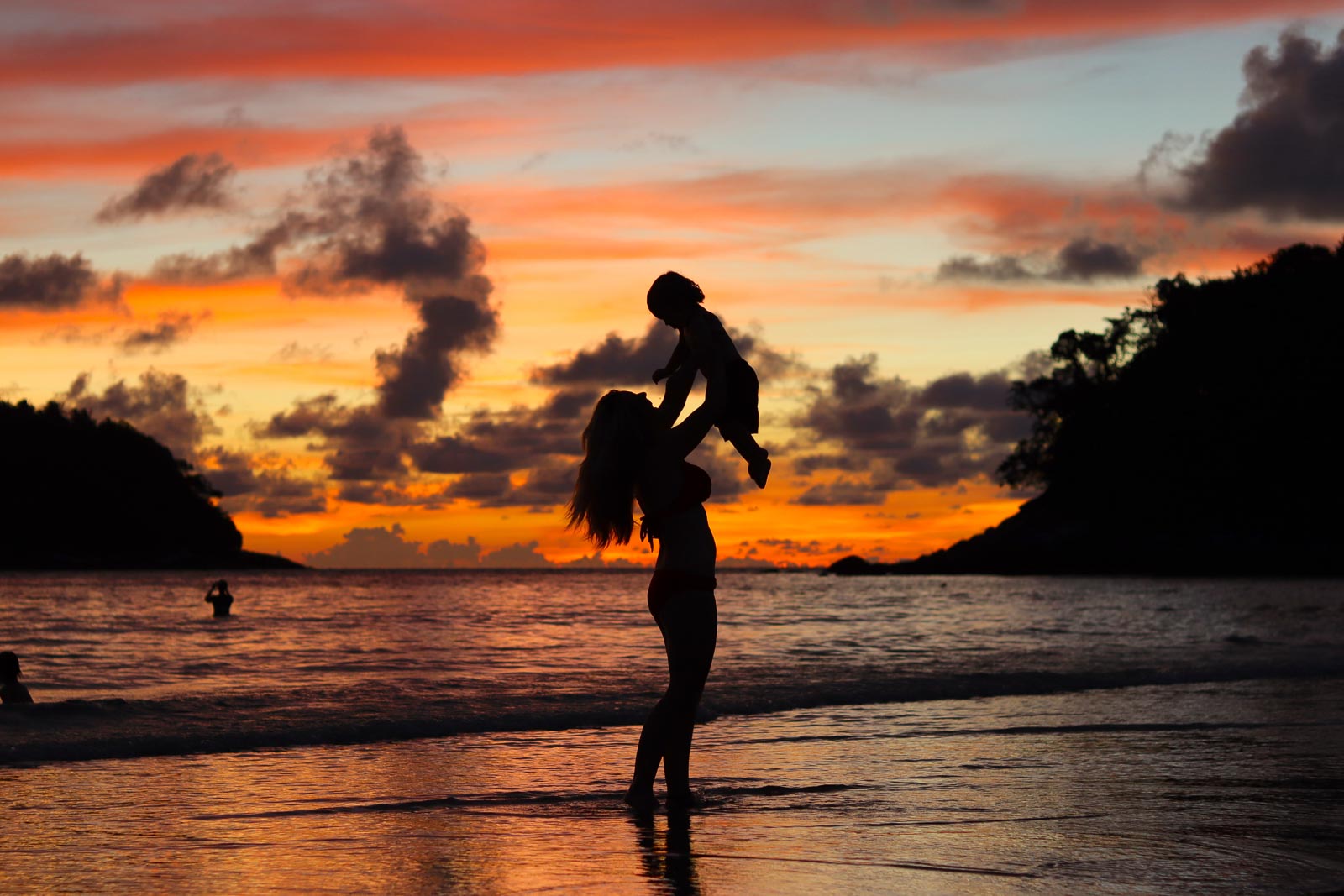 woman holding a baby above her head with an unreal orange andpinkish sunset on the beach