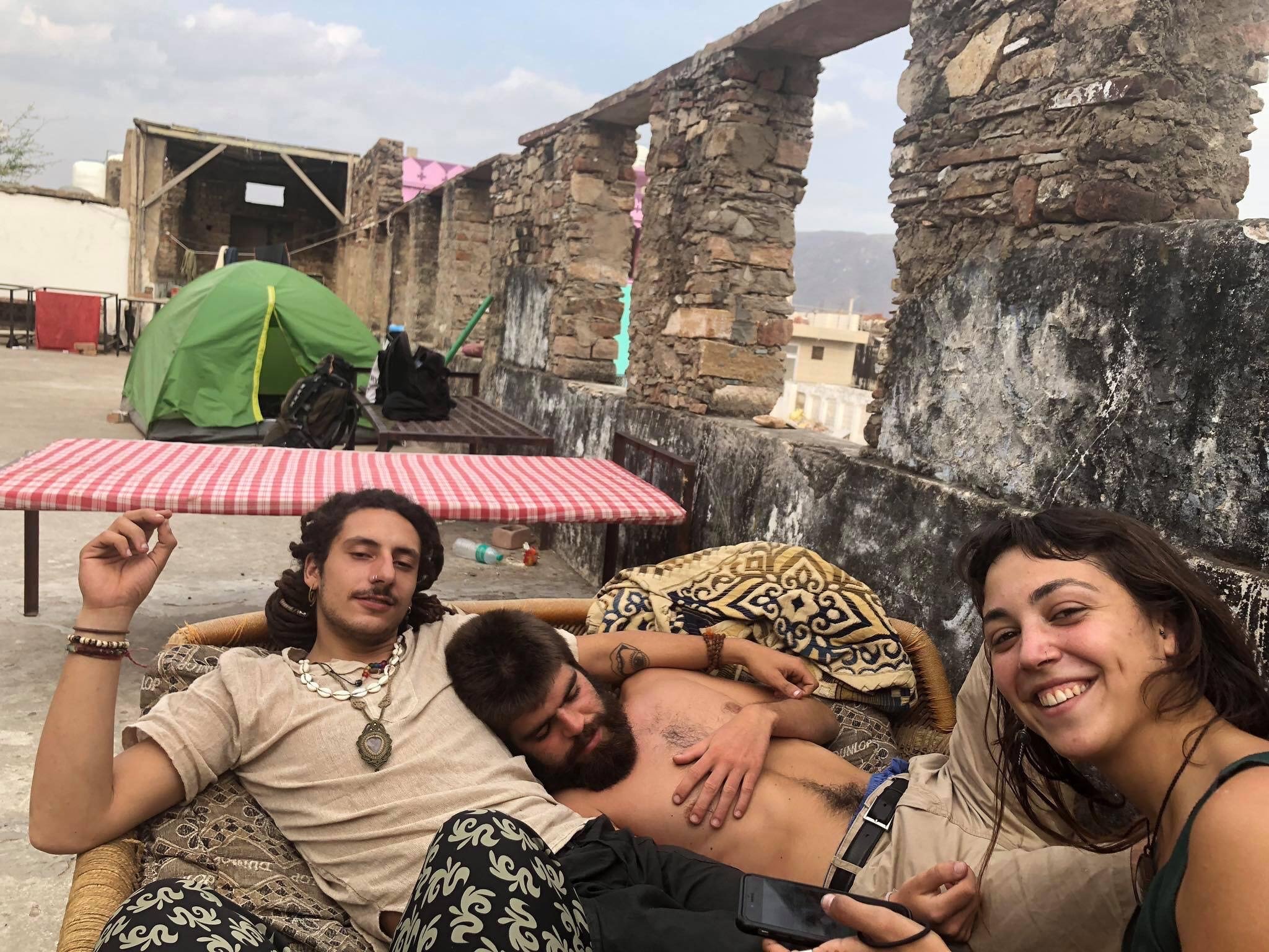 three people lounging on a charpai hammock on a rooftop while staying at a hostel in india
