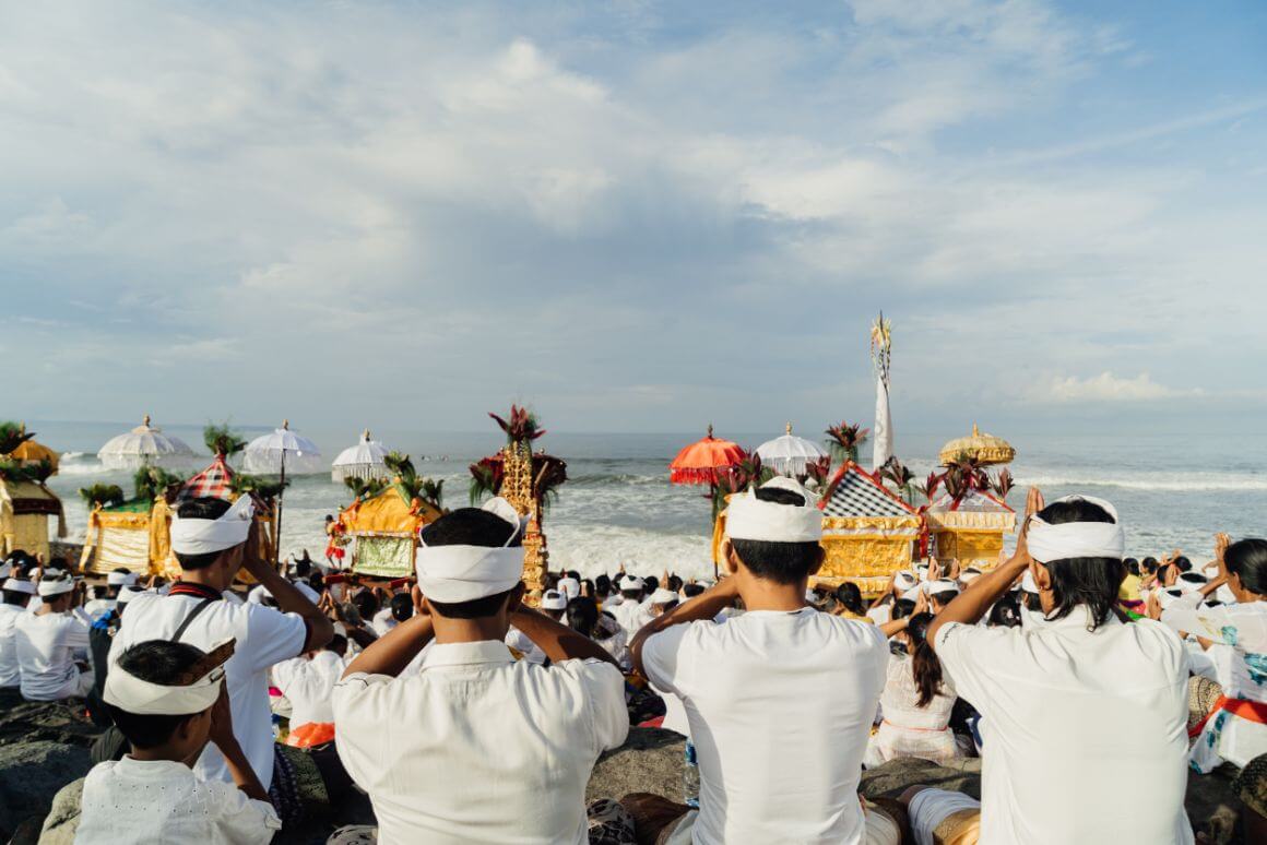 A group of Balinese citizens in traditional white clothing and headbands sitting on a beach facing the ocean with several ornate ceremonial structures and umbrellas in front of them.