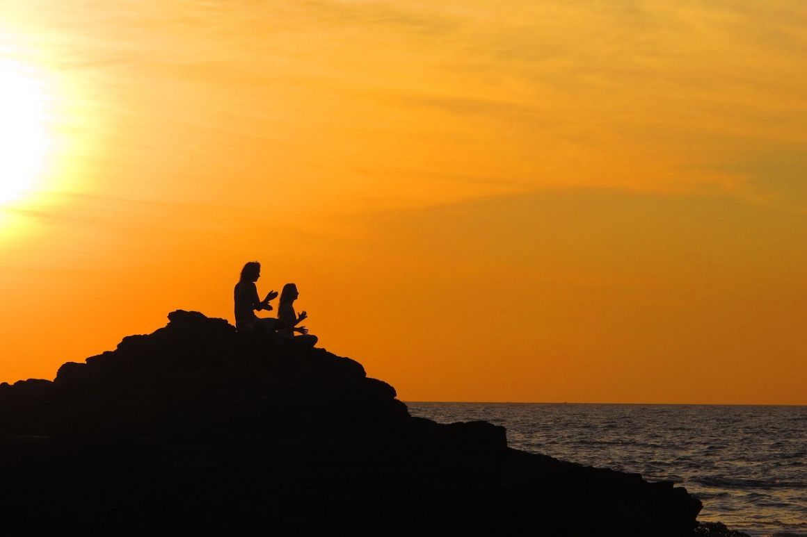 two people doing yoga on a rock alongside the ocean during an incredible orange sunset in goa india
