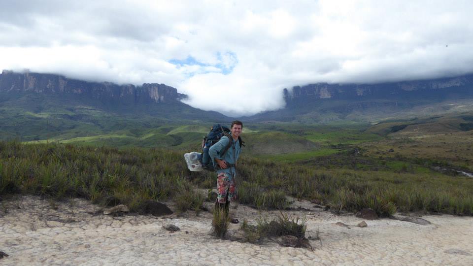 man carrying a. large backpack while standing in front of a green mountain range in venezuela
