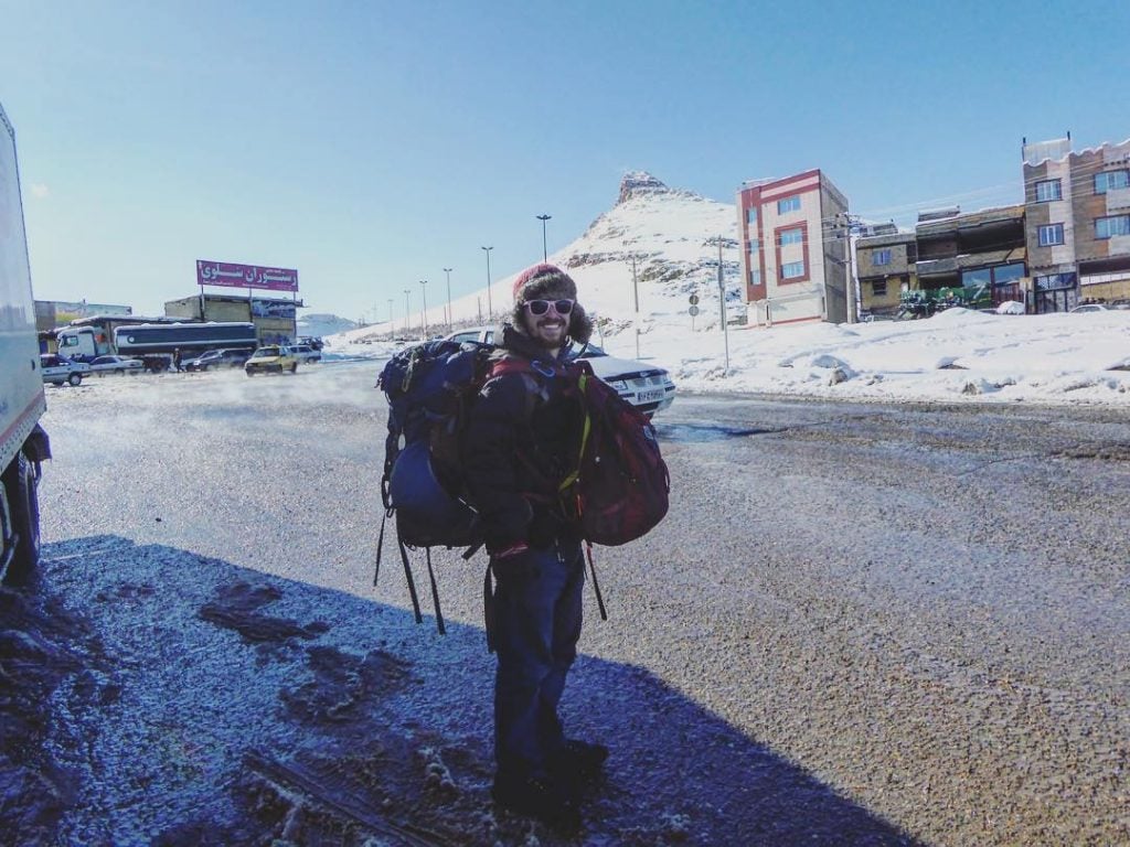 backpacker holding a backpack waits on the side of the road for his next hitchhiking ride