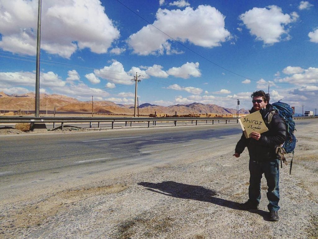 man holding up a cardboard sign on the side of the road while hitchhiking in iran