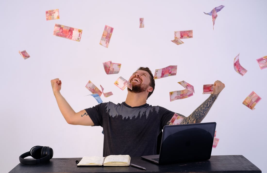 excited man / will sitting at a desk with a journal and a laptop with money notes in the air and raining down on him