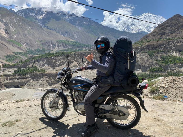 A man on a motorcycle/ motorbike with a large backpack and camping gear attached doing a shaka sign whilst stopping for a break whilst riding through the mountains. In the background is a valley with several mountains, some covered in snow