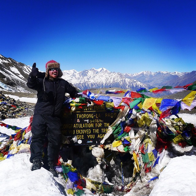 Will at the top of a mountain in Pakistan with the jagged snow covered peaks of the Himalayas behind him and prayer flags wrapped around a sign.