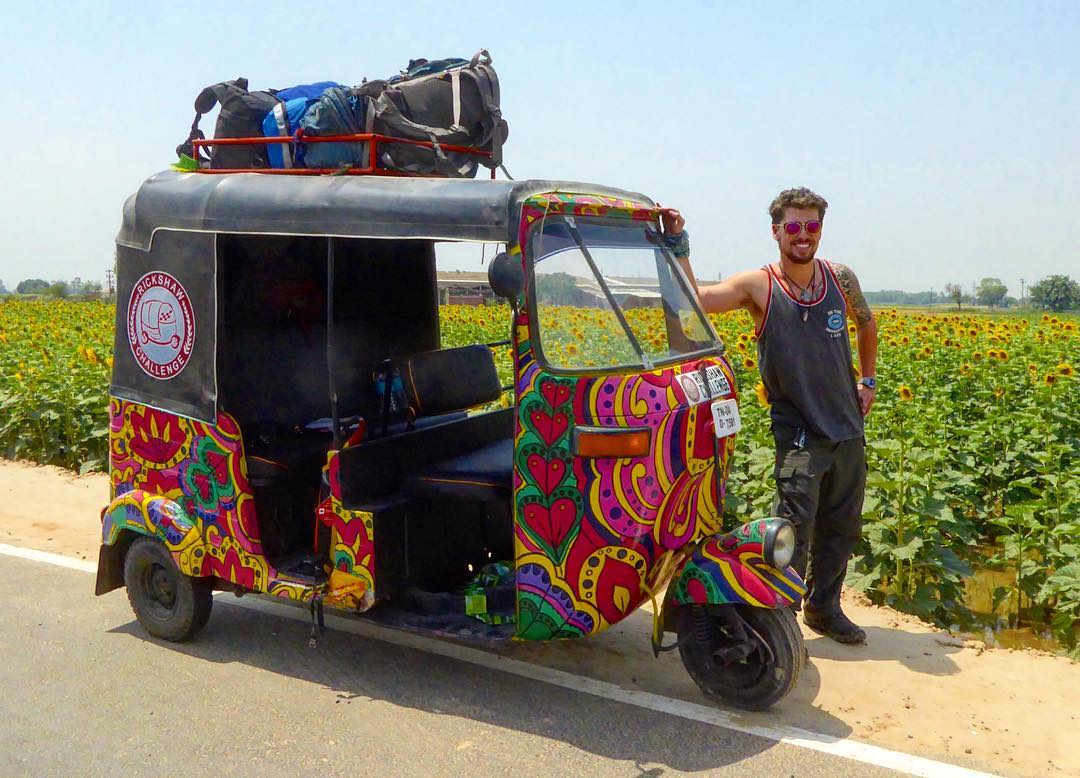 Will leaning on a colourful rickshaw/ tuk tuk in India with a field of sunflowers behind him and a pile of backpacks on the roof