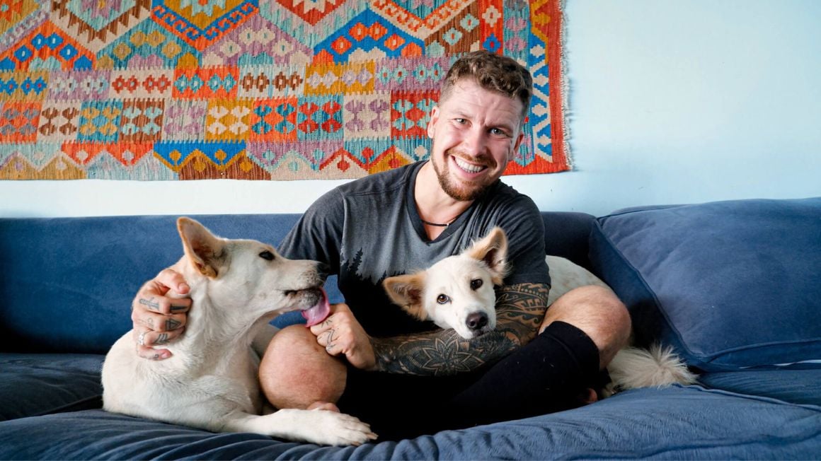man sitting on a blue couch with two white dogs anda colorful tapestry on the white wall behind him