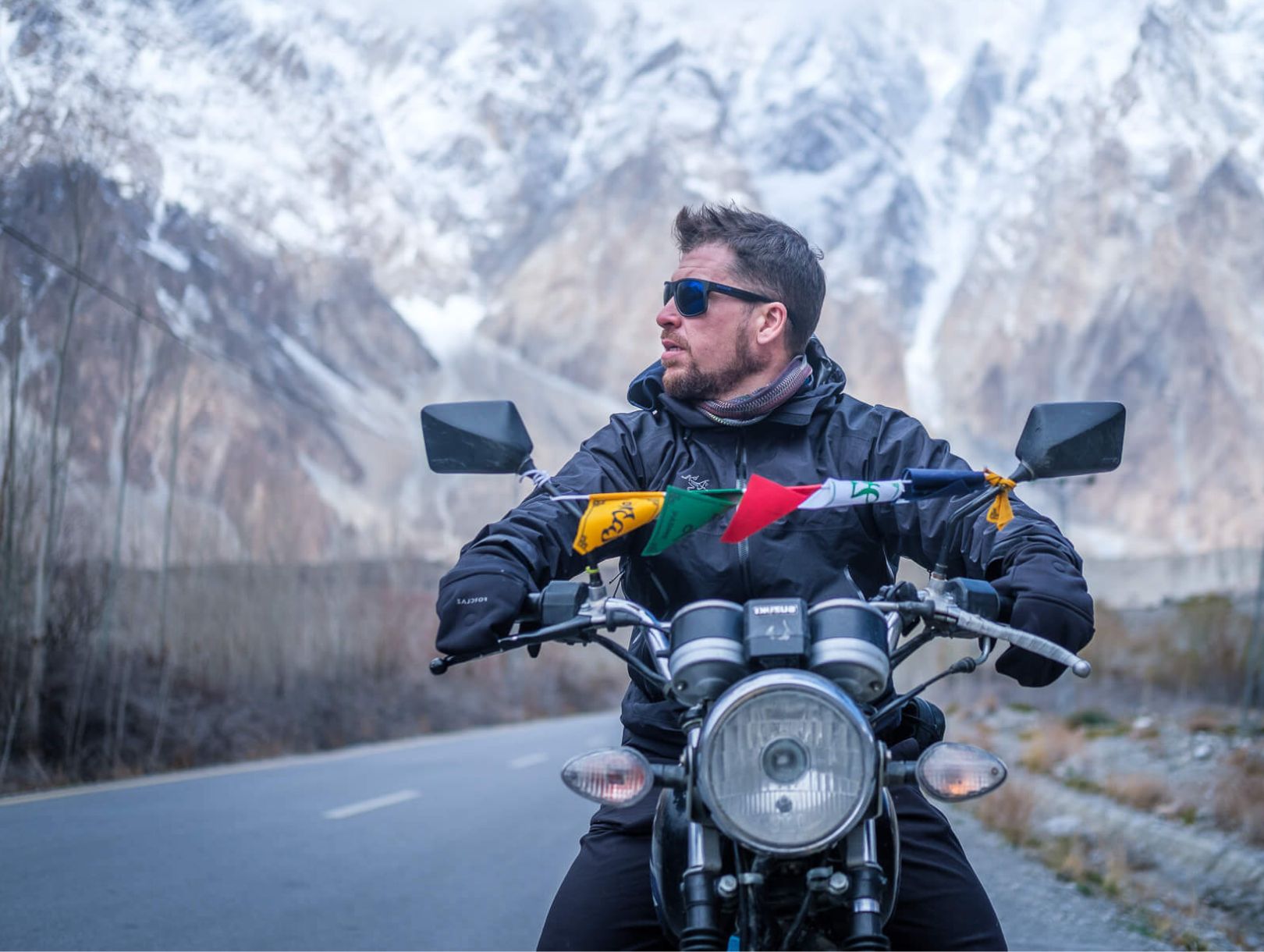 will sitting on a Pakistani motorcycle strung with tibetan prayer flags with massive snow-covered peaks behind him