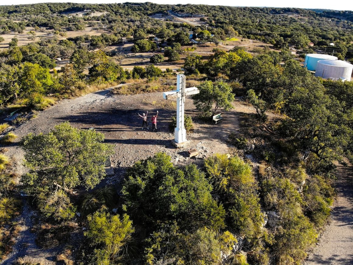 A couple of people standing next to a giant white cross on top of Cedar hill surrounded by trees 