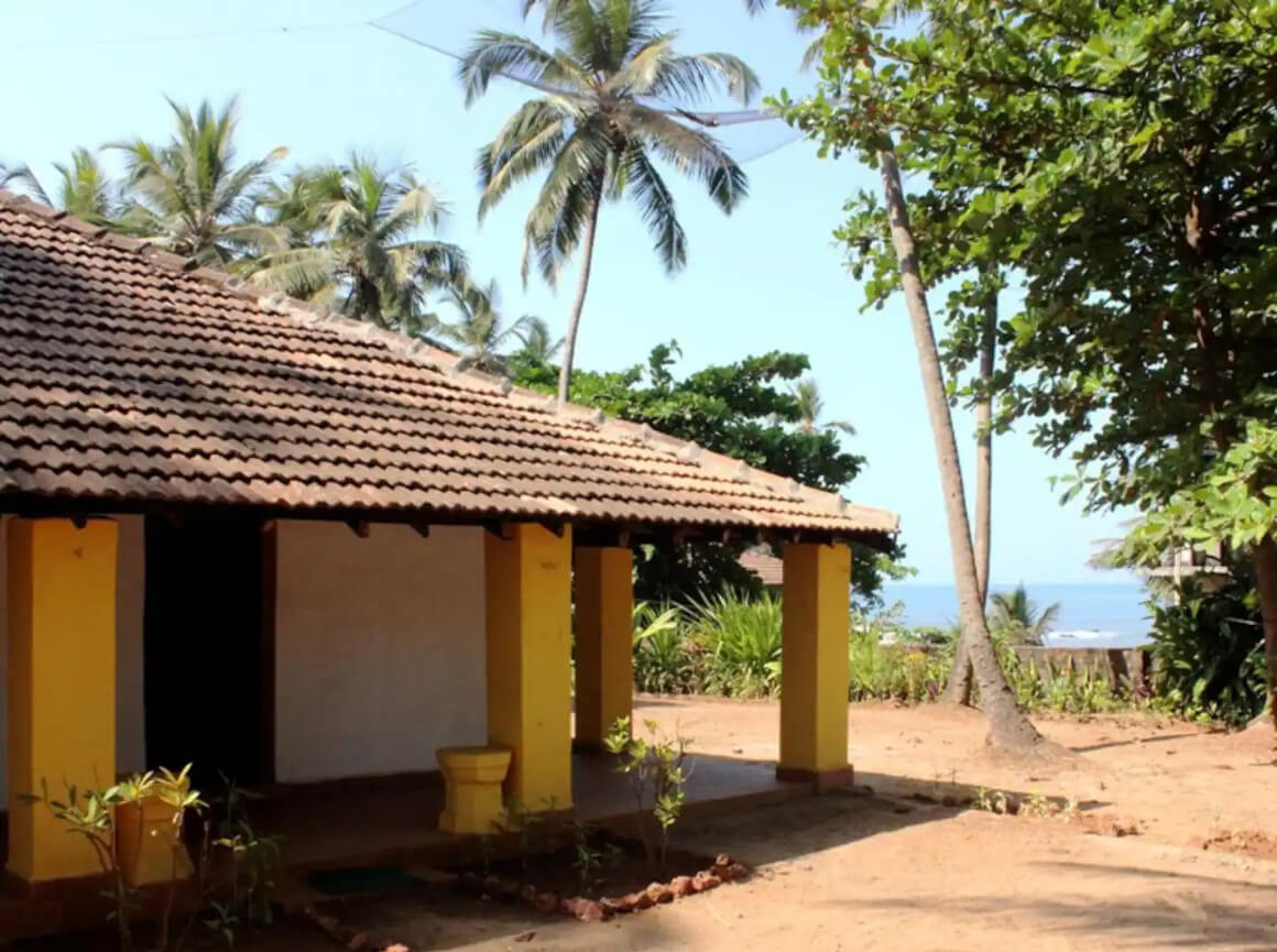 simple yellow Goan Beach House with a thatched roof surrounded by palm trees