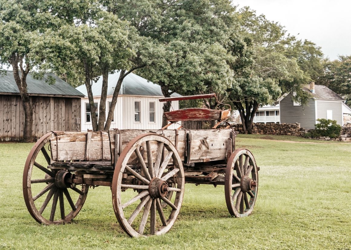 A wooden wagon sitting in a field with barns in the background surrounded by nature