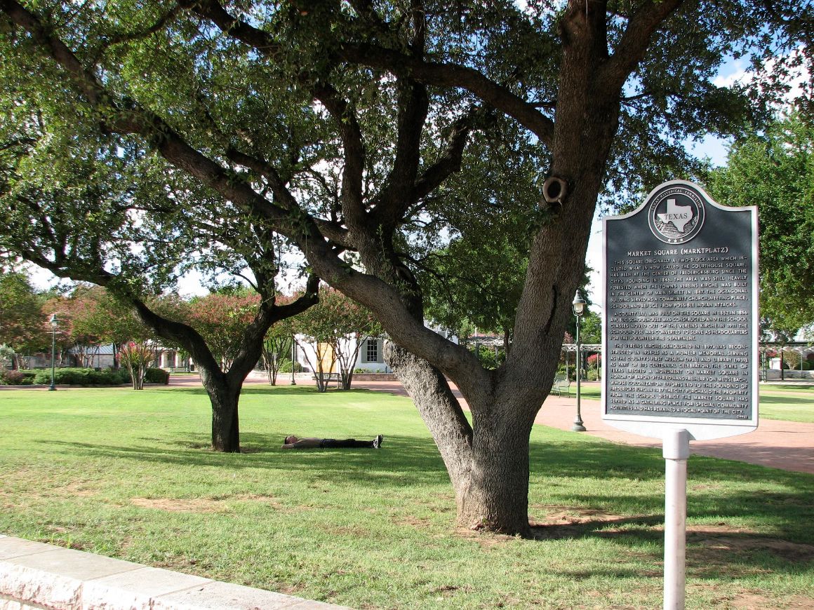 A person laying on the ground under a tree in Marktplatz Fredericksburg.