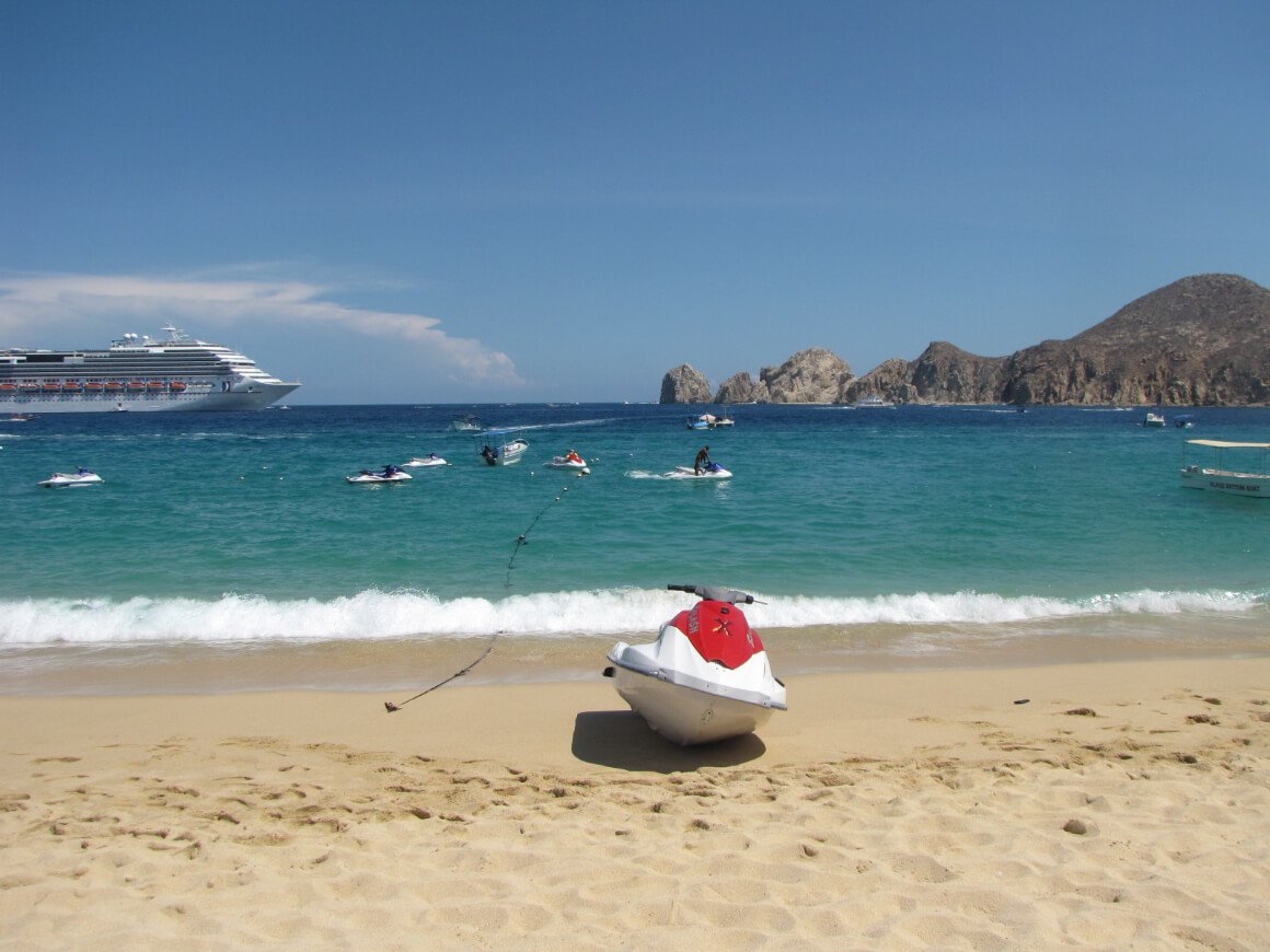 A landscape view of Medano Beach featuring a jet ski resting on the shore, a boat and rock formation in the middle of the ocean