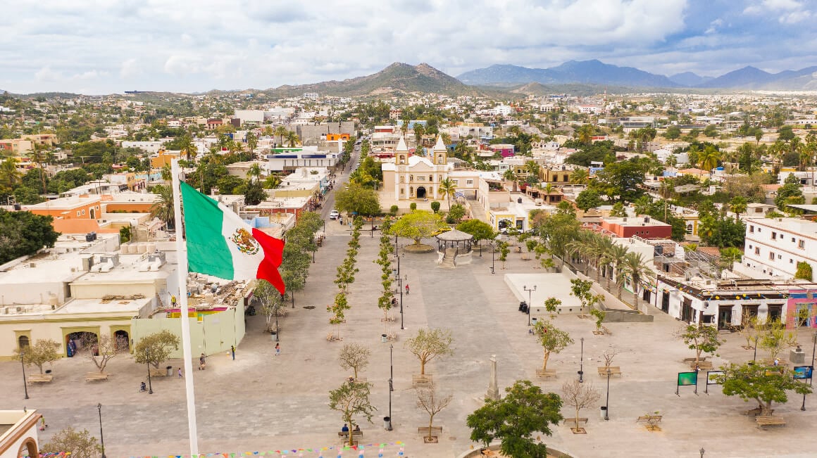 San Jose Del Cabo cityscape with Mexico's flag, framed by lush trees and mountains in the background