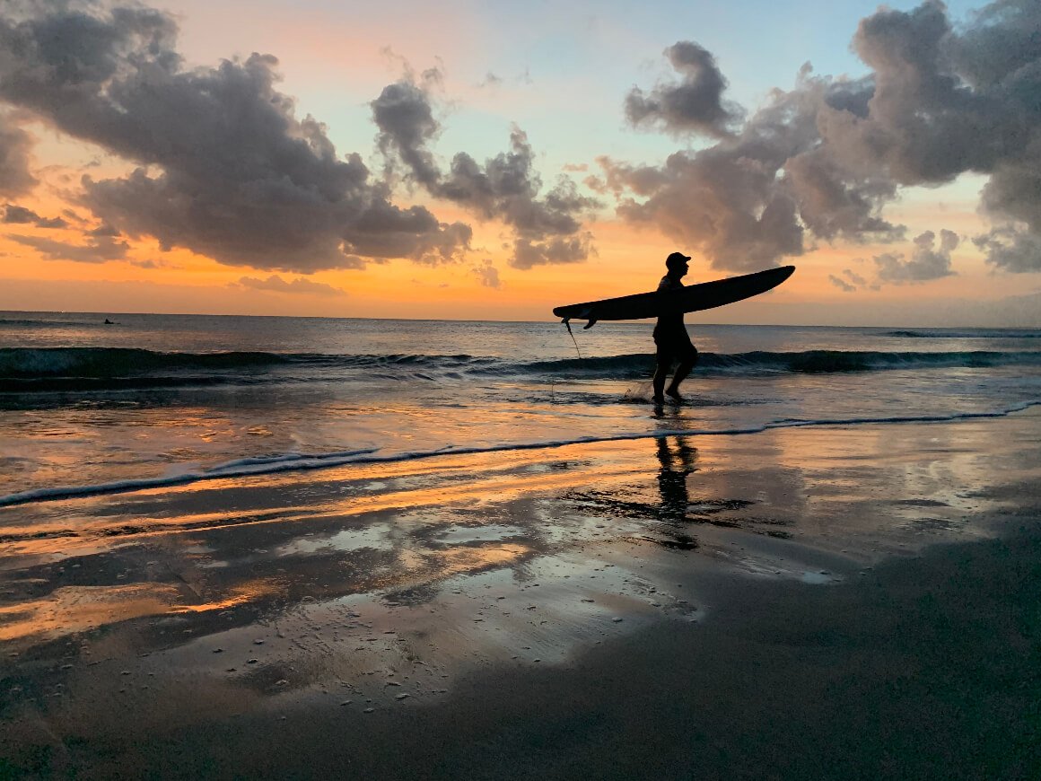 person walking out of the sea with a surfboard at sunset