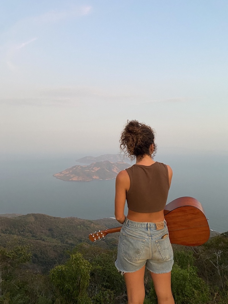 Girl stands on top of volcano with guitar and looks out over view of El Salvador.
