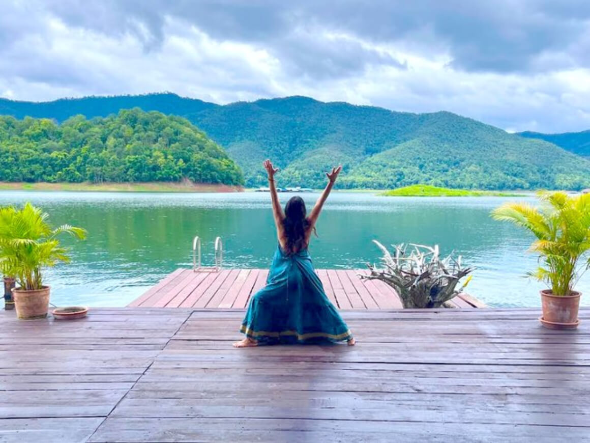 A woman adorned in a blue hippy dress immersed in a yoga pose, gazing at a majestic river surrounded by lush greenery.