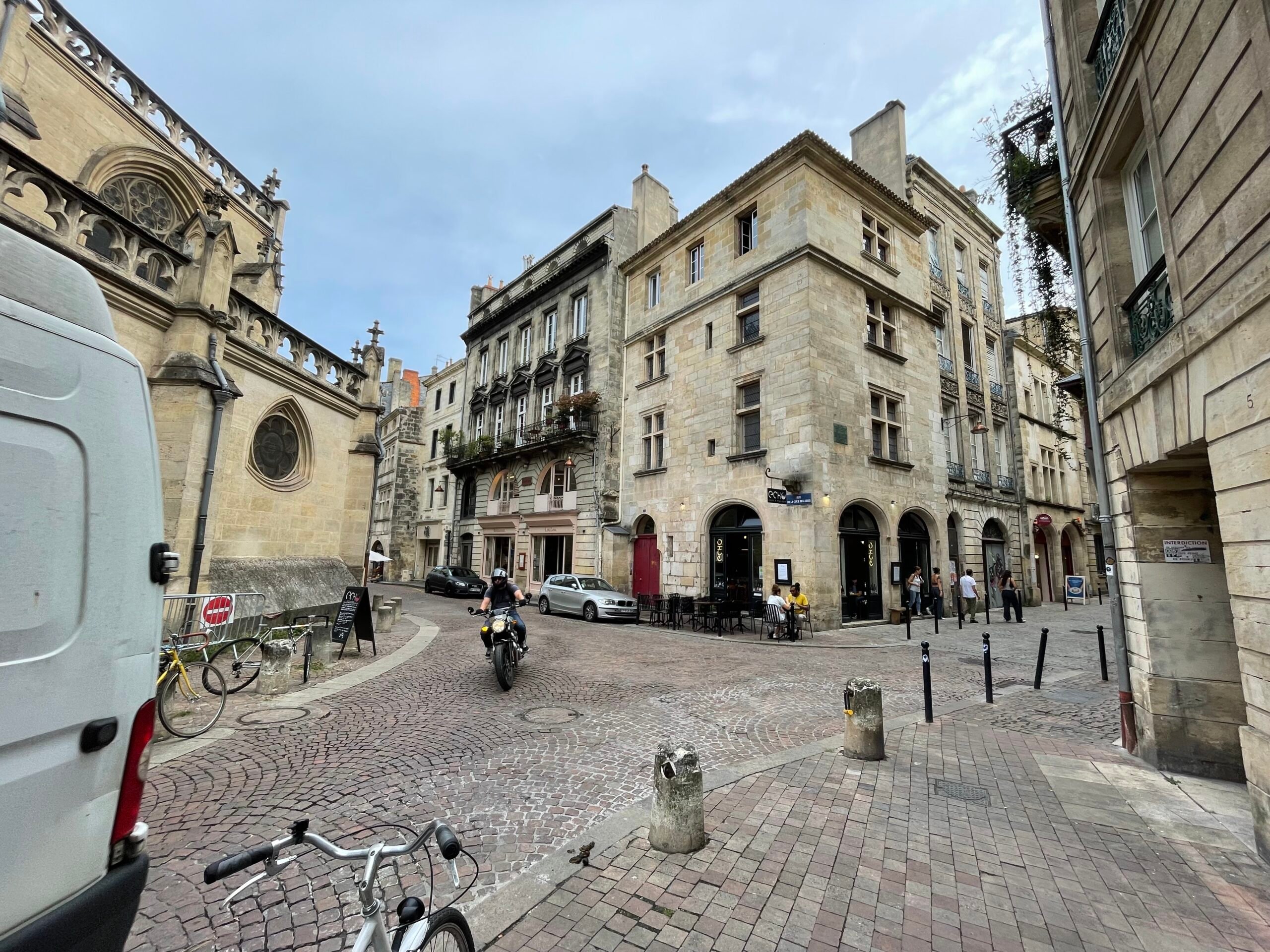 A view of Bordeaux's narrow streets with old stone buildings, featuring a person riding a motorcycle and people strolling.
