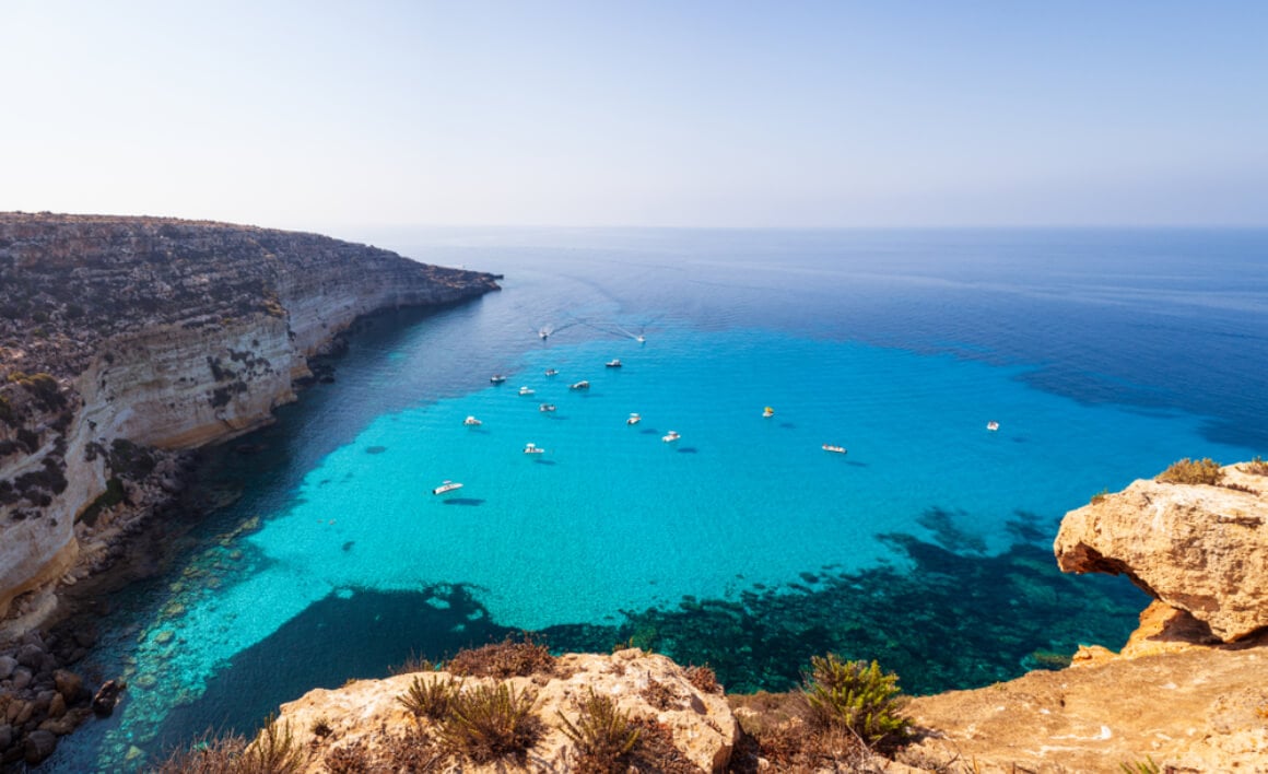 Boats floating in turquoise waters surrounded by cliffs in Lampadusa, Italy