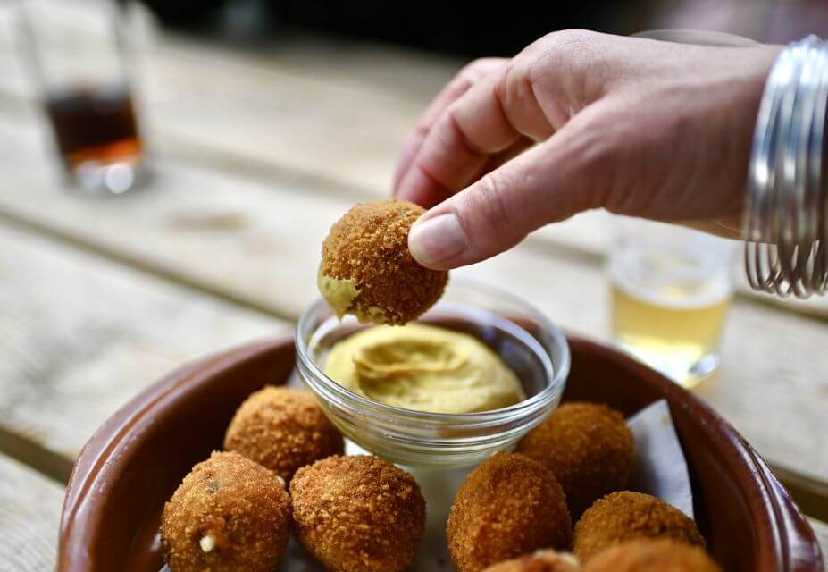 A person's hand dipping a fried ball into some mustard