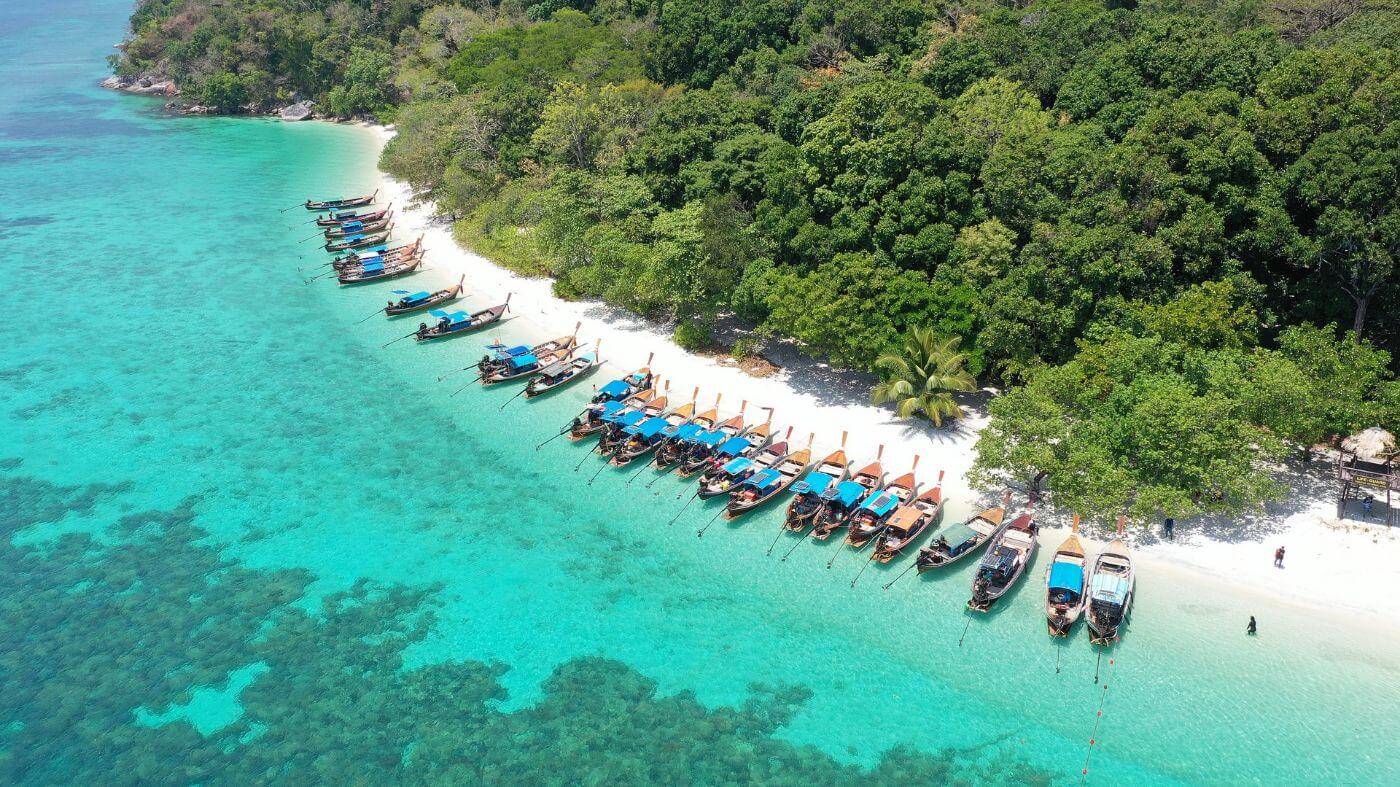 A landscape view of a group of boats lined at the shore of Koh Lipe, Thailand facing a lush forest 