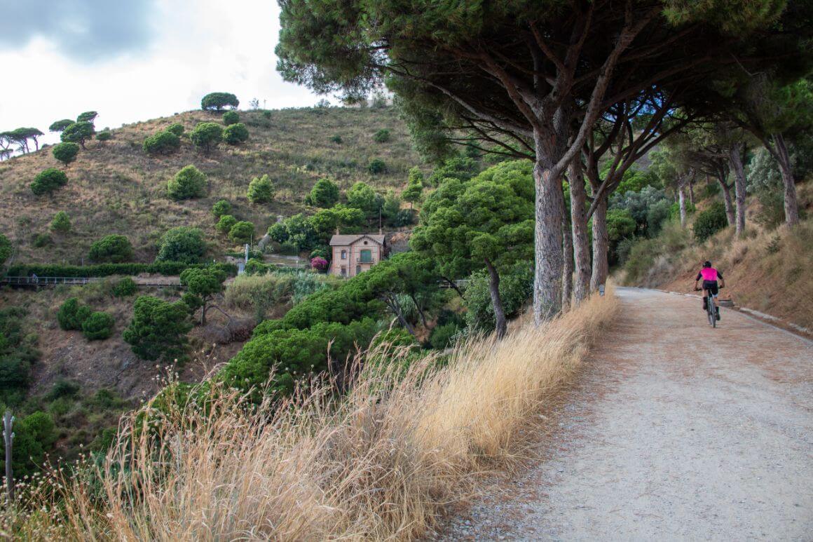 A person riding a bicycle down a dirt road with lush trees and a house in the background from La Carretera de les Aigües, Barcelona