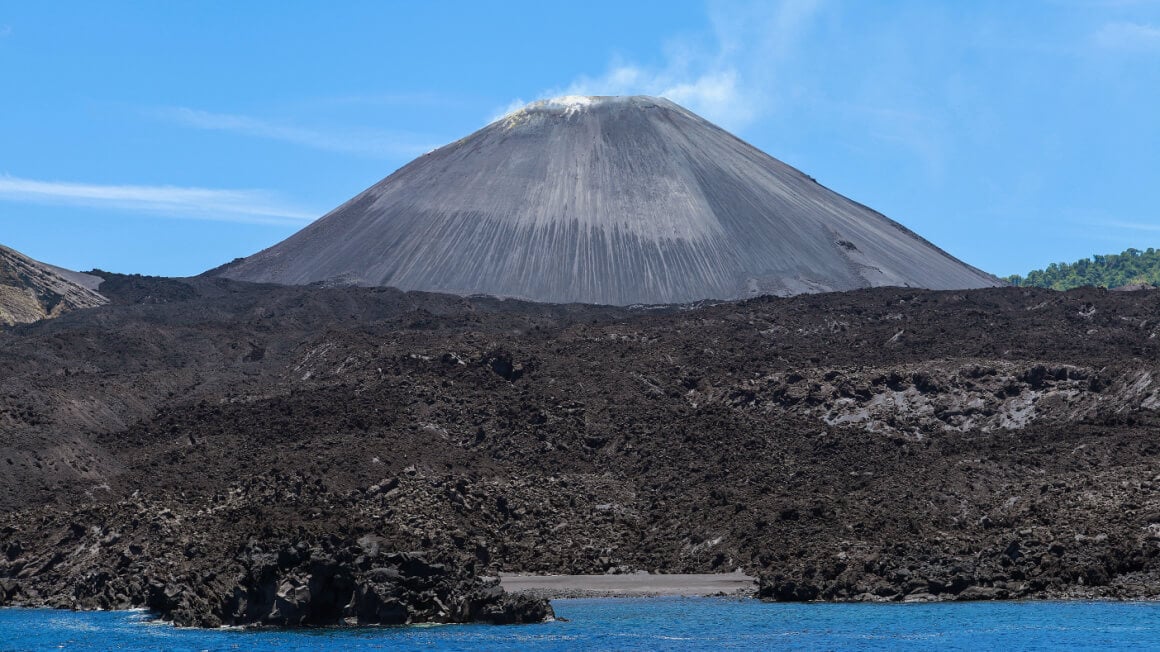 a volcano rising up above a ton of black ash next to the ocean on an island in India