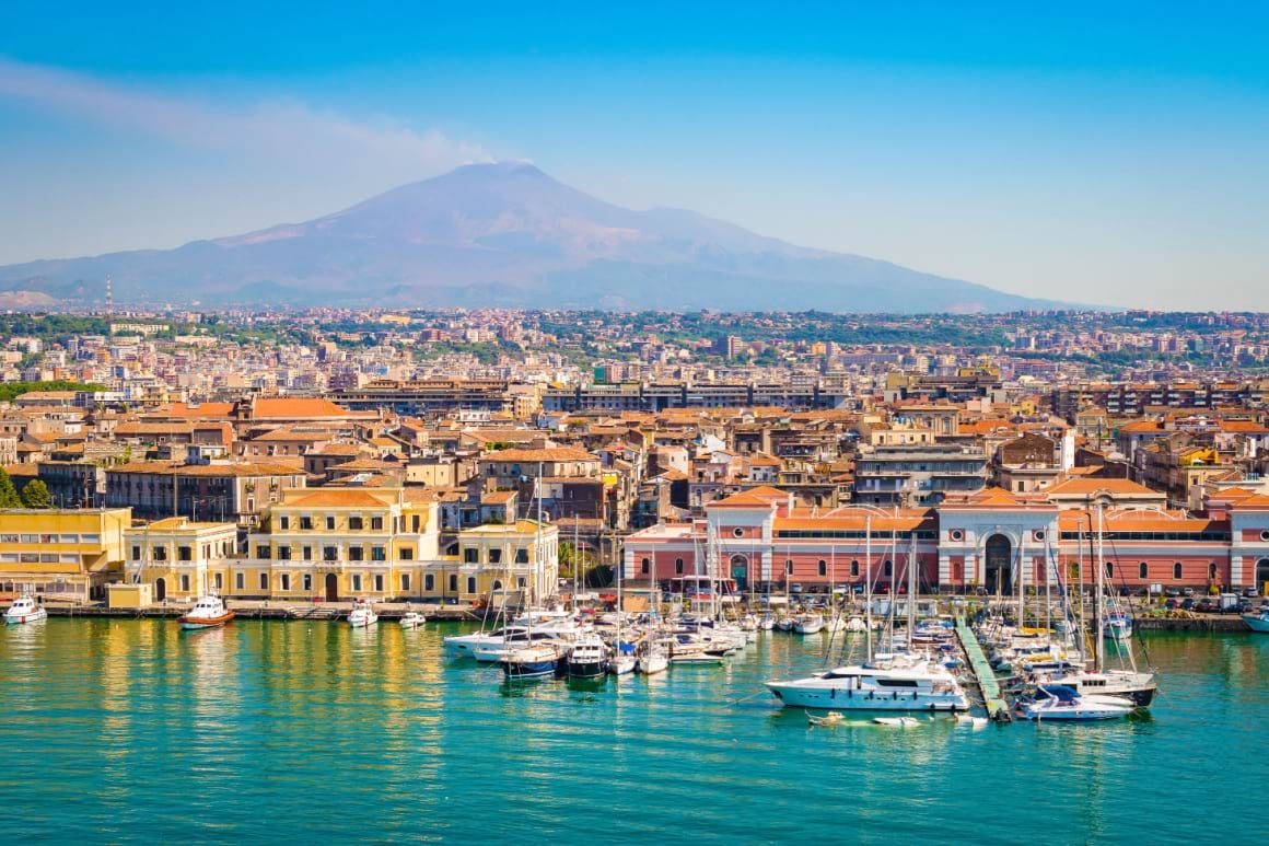 colorful building along the water and a huge mountain peak rising up in the distance in catania