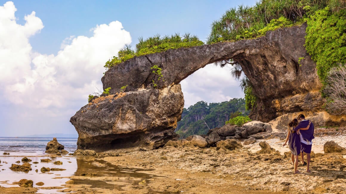 couple walking on a beach in India toward a bridge-shaped rock formation that's covered in lush greenery
