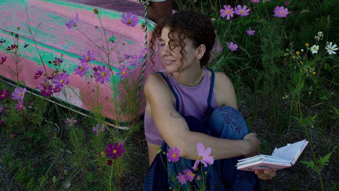 Audy wearing a purple top holding a book looking around surrounded by purple flowers
