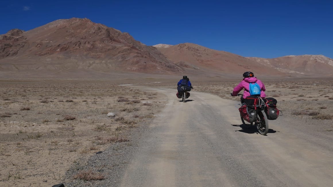 two cyclists with fully loaded bags cycling on a dirt road in a reddish mountain landscape while on an offbeat adventure in tajikistan